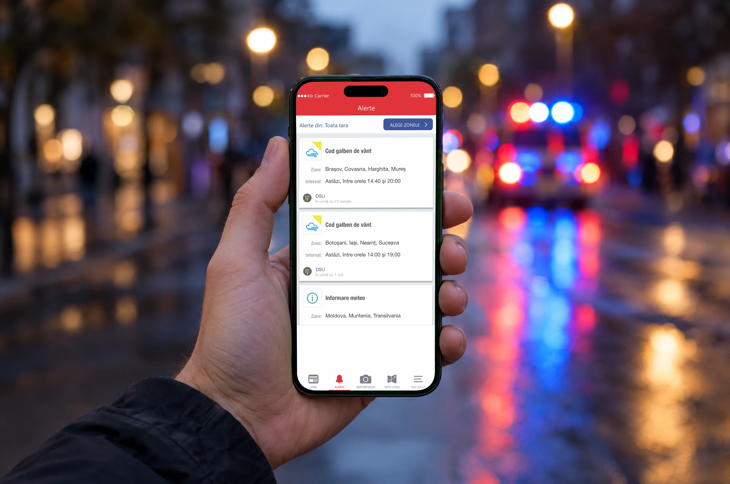 The DSU app's alerts on a phone held in one hand, in a wet street at dusk with an emergency vehicle's lights far behind