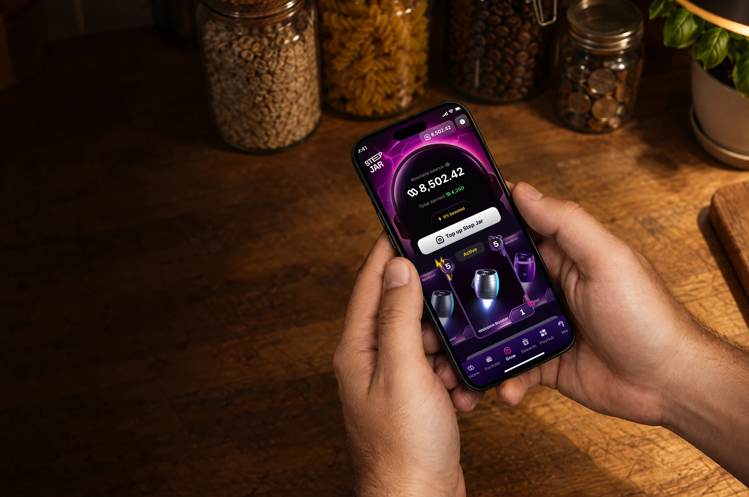 Seen from above, two hands hold a phone over a kitchen counter, glass jars at its back; on the screen, Sweat Wallet’s Step Jar: a boostable balance of 8,502.42 and a Top up Step Jar button