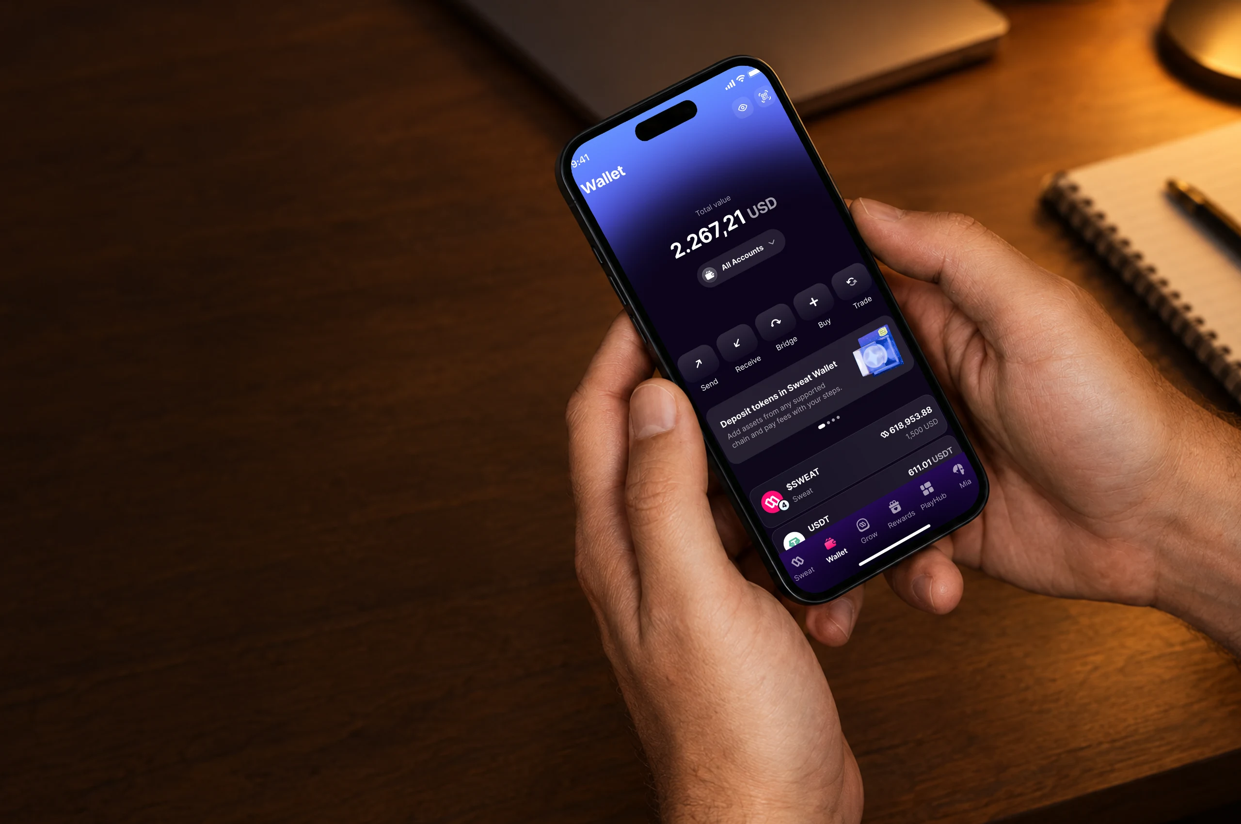 Seen from above, two hands hold a phone over a wooden desk in the evening, a notebook and a pen beside it; on the screen, Sweat Wallet’s Wallet page: a total value of 2.267,21 USD, and Send, Receive, Bridge, Buy and Trade