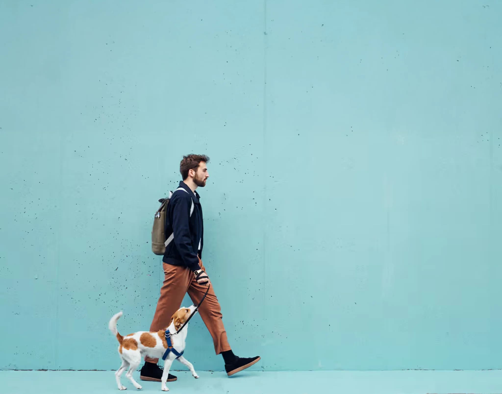 A man walks his dog along a turquoise wall, a backpack on his shoulders